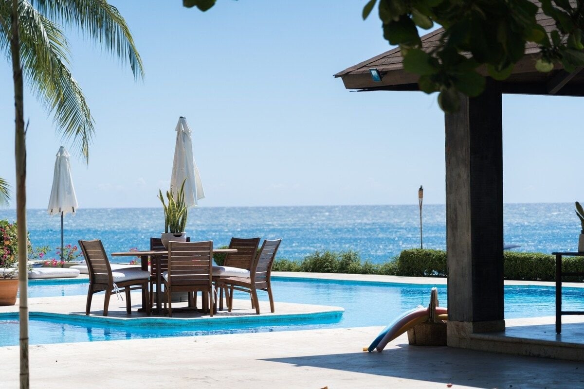 The outdoor area features a spacious pool with crystal clear water, surrounded by sun loungers and wooden dining furniture. Umbrellas provide shade, while the ocean is visible in the background, with a blue sky above enhancing the seaside ambiance.