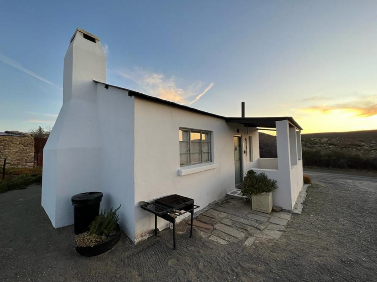 A quaint, single-story building is shown, featuring a white exterior and a flat roof. A chimney extends from one side. Accessibility is provided by a stone pathway leading to the entrance, while a braai area is positioned outdoors next to a small planter.