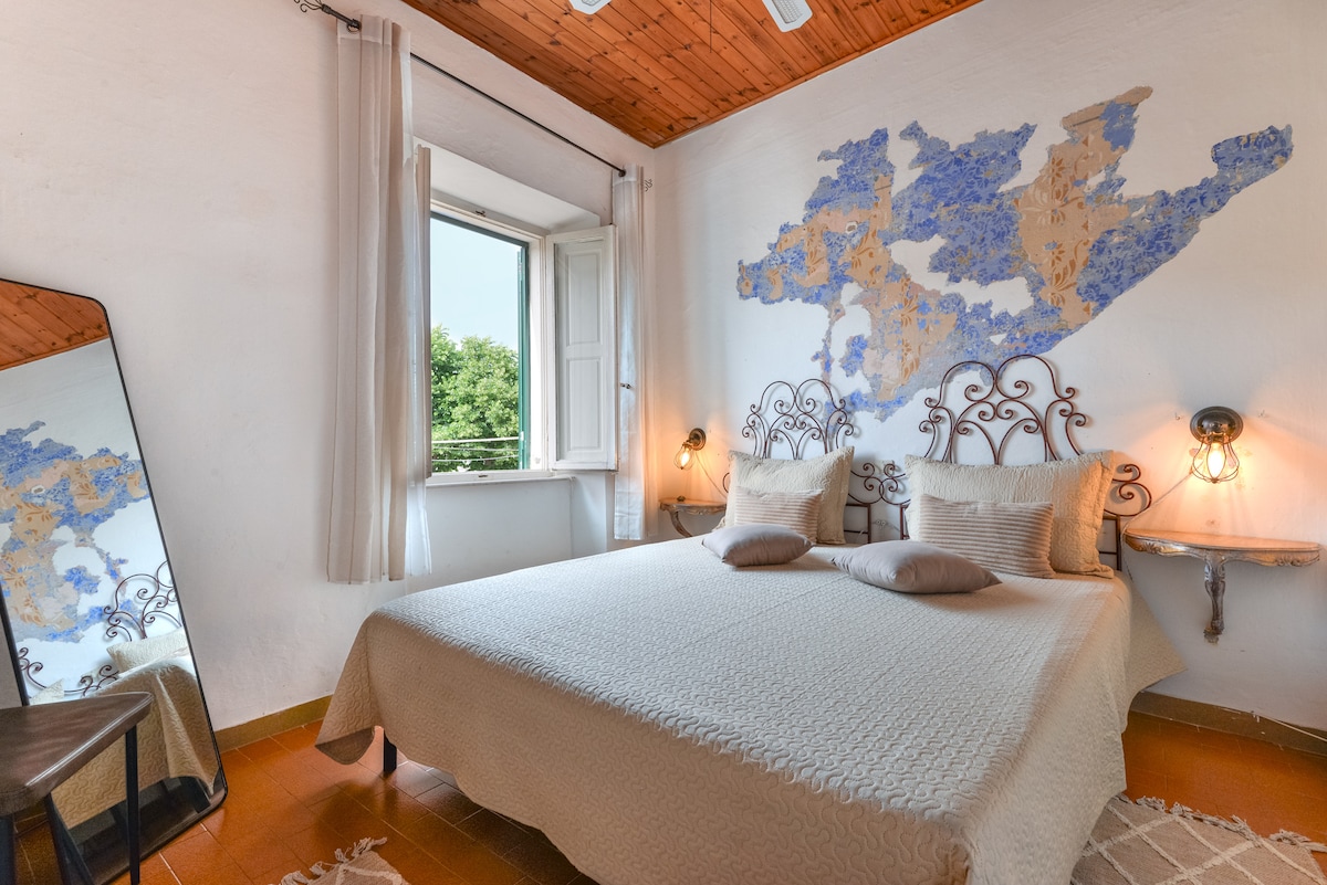 A serene bedroom is featured, showcasing a spacious bed adorned with neutral linens and two cushions. A decorative wall mural adds character. Natural light enters through an open window, while a full-length mirror reflects the room's ambiance. A small side table is positioned beside the bed.