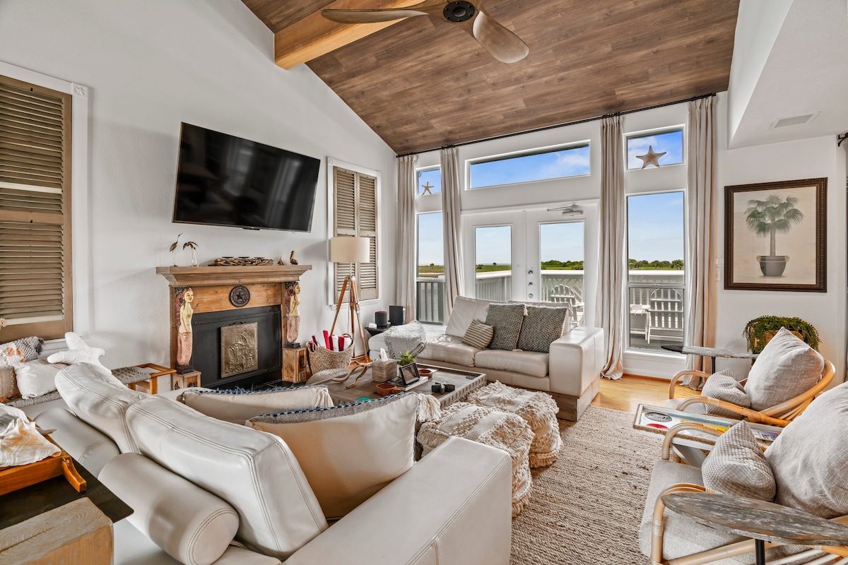 A bright and spacious living area features a comfortable arrangement of white sofas and assorted chairs. Large windows provide views of the natural surroundings, while a wooden ceiling adds warmth. A fireplace is framed by a decorative mantle, enhancing the coastal decor.