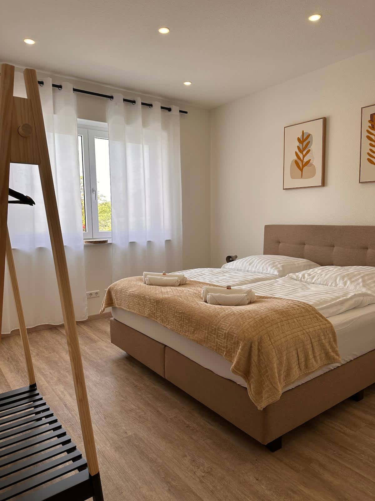 A well-lit bedroom features a spacious bed with crisp white linens and a warm textured blanket. Two decorative wall art pieces are displayed above the bed. Sheer white curtains frame the window, allowing natural light to fill the room. A wooden clothing rack is positioned nearby.