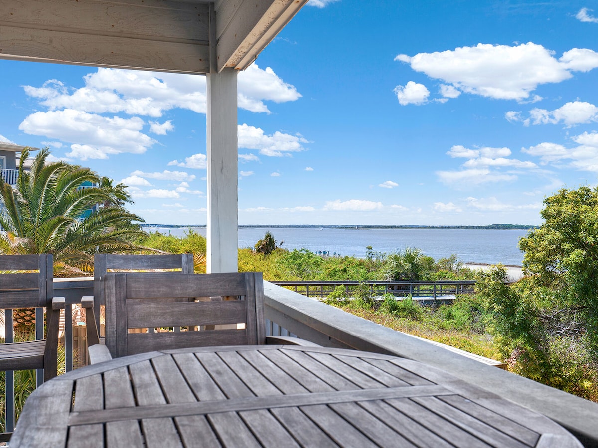 An outdoor balcony features a wooden dining table and chairs, positioned to provide views of the nearby water and lush greenery. Palm trees can be seen in the foreground, with a clear blue sky and soft clouds enhancing the peaceful atmosphere.