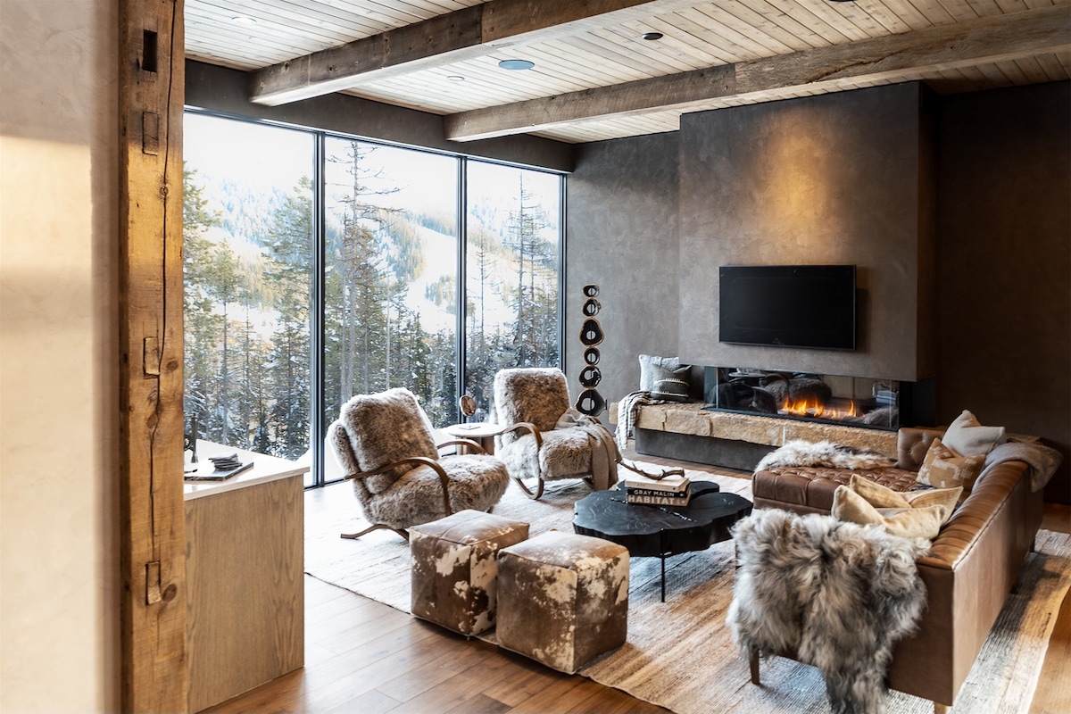 The living room showcases a modern design with large floor-to-ceiling windows offering serene views of the surrounding trees. A leather sofa and plush chairs are arranged around a low coffee table, complemented by neutral tones and cozy textures, creating a warm space for relaxation.