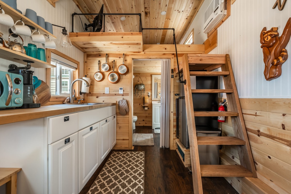 An open-concept kitchen and living area showcases wood paneling and modern appliances. A staircase leads to a lofted space, while cooking essentials are neatly arranged on shelves. Natural light filters through windows, illuminating the space and highlighting the cozy aesthetic.