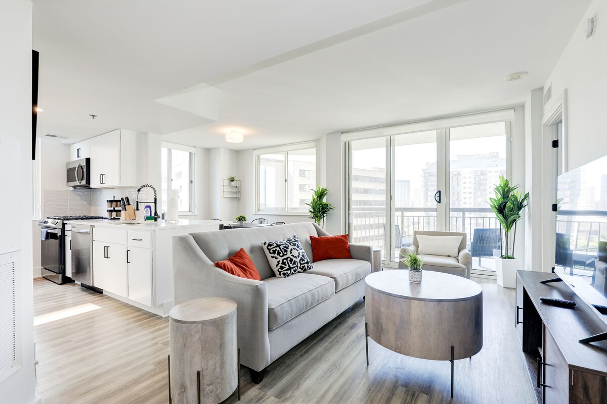 A bright and airy living space featuring a comfortable sectional couch adorned with colorful throw pillows. Large windows allow natural light to flood in, while a round coffee table sits centrally. A modern kitchen is visible in the background, complete with sleek appliances and bar seating.
