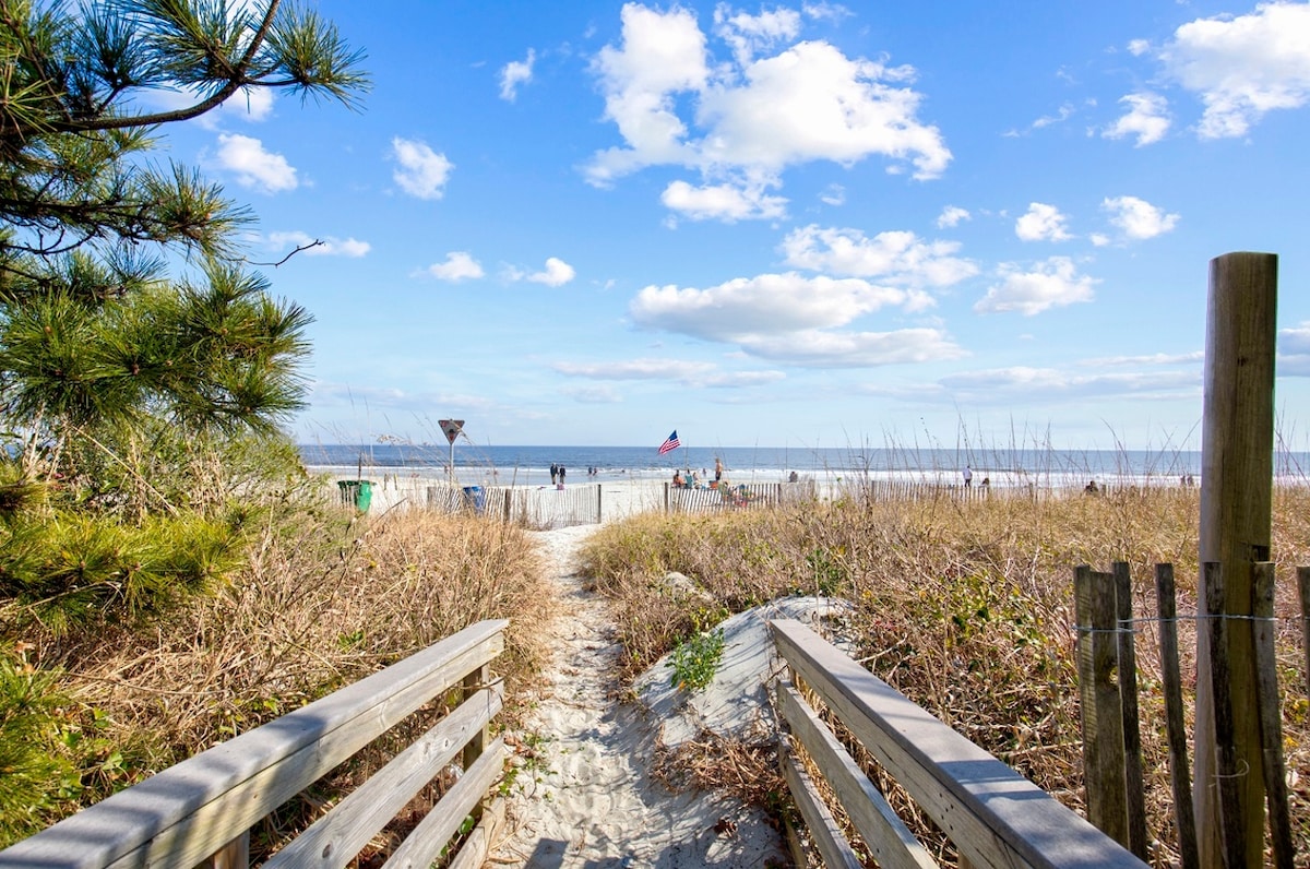 A wooden pathway leads through sandy terrain towards the beach, framed by tall grasses and small trees. The ocean is visible in the distance, with gentle waves and a clear sky dotted with clouds. A few beachgoers are seen enjoying the shore.