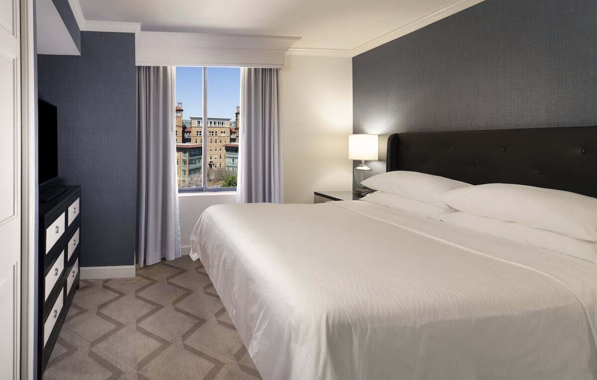 A spacious hotel room features a king bed with crisp white linens, complemented by a sleek black headboard. A window provides natural light and views of the city. A dresser stands against the wall, while soft grey curtains frame the window.