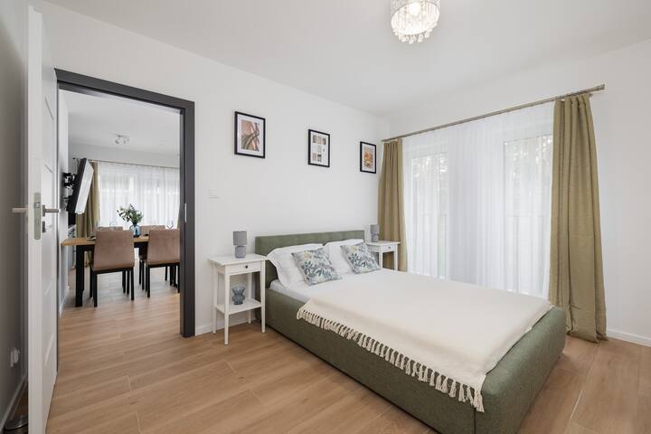 A bedroom with a large bed and simple decor. Light-colored curtains frame the windows, giving the room a fresh and airy feel.