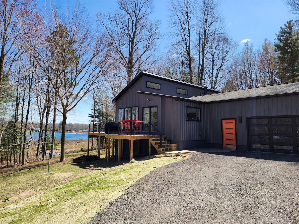 A contemporary home is set on a grassy area with a gravel driveway leading to a garage. Large windows offer views of the lake, while a red railing surrounds the wrap-around deck. Surrounding trees provide a natural backdrop, creating a serene setting.