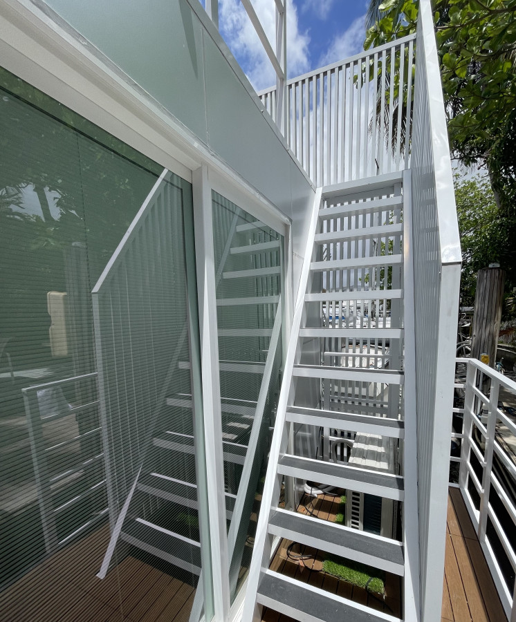 A set of white exterior stairs leads to an upper deck, positioned beside large glass windows. Sunlight reflects off the sleek railing and the wooden deck below, framed by surrounding greenery.