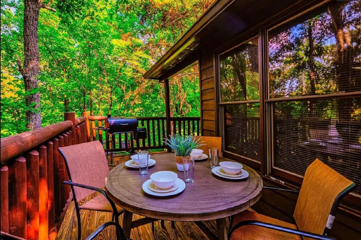 A deck area is presented with a round wooden table surrounded by four cushioned chairs. The table is set with plates and glasses, while lush green trees provide a natural backdrop and the fading sun casts a warm glow through large windows.