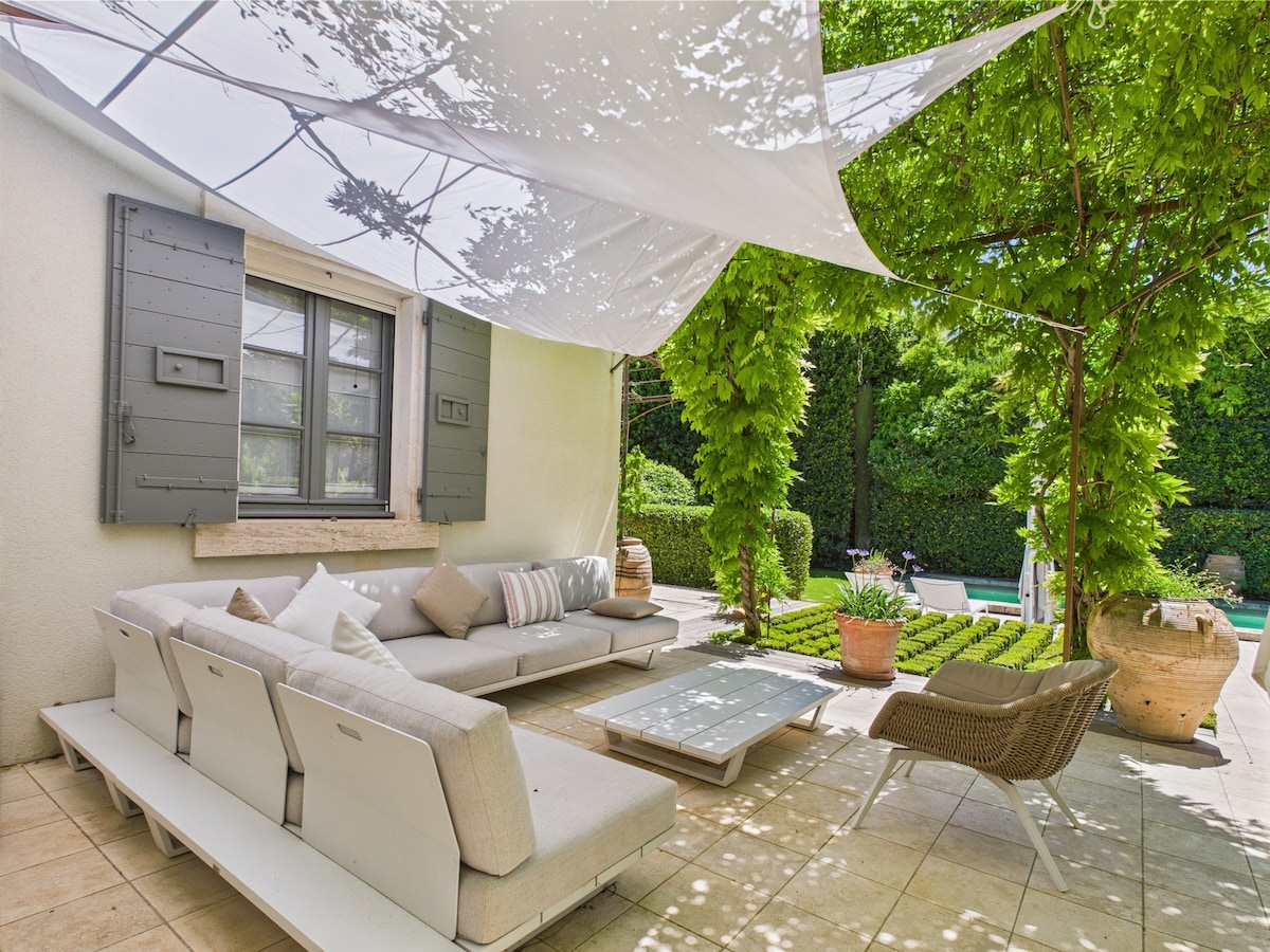 A shaded terrace area features a spacious L-shaped seating arrangement, complemented by a low coffee table. Lush greenery frames the space, creating a serene atmosphere. A glimpse of the swimming pool is visible in the background, enhancing the setting's tranquil appeal.
