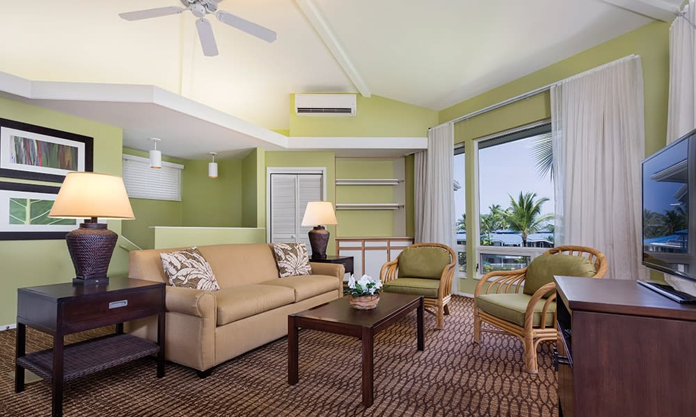 The living area is characterized by light green walls and a high ceiling, complemented by a large window offering views of palm trees. A comfortable sofa, two chairs, and a wooden coffee table create a relaxed seating arrangement, while a flat-screen TV is positioned nearby.