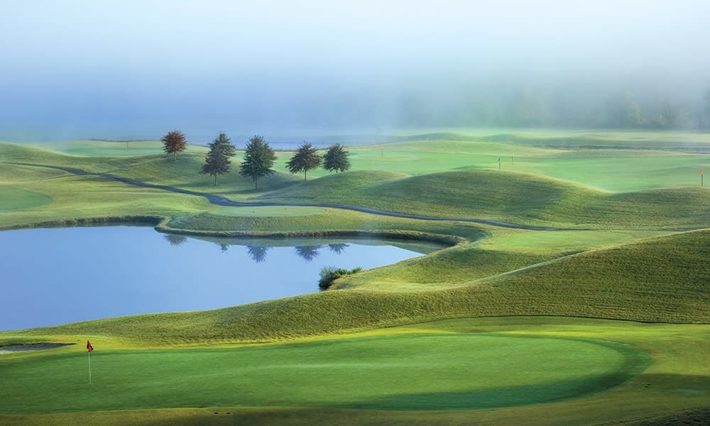A lush green golf course is seen, featuring rolling hills and a serene pond reflecting the landscape. Gentle fog envelops the area, adding a tranquil atmosphere. Several trees are positioned along the edge of the course, complementing the peaceful surroundings.
