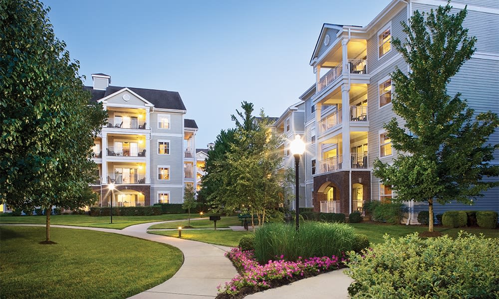 The resort grounds are lined with well-maintained pathways, surrounded by lush greenery and flowering plants. Multiple building entrances are visible, with warm lights glowing from the balconies. A softly lit lamppost adds to the inviting atmosphere of the outdoor space.