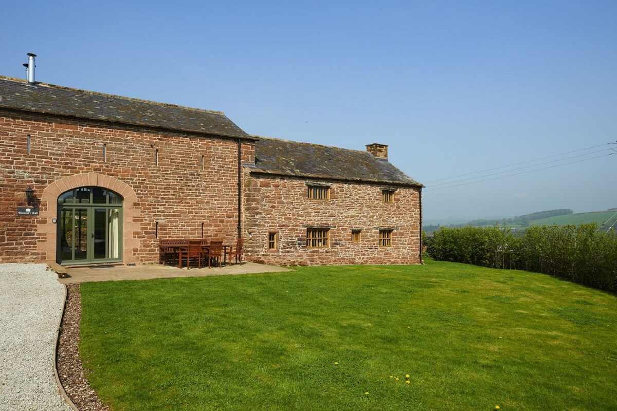 A rustic stone building is set against a clear blue sky, featuring large windows that provide ample natural light. A well-maintained lawn surrounds the property, complemented by a wooden dining table and chairs positioned on a patio area, ideal for outdoor gatherings.