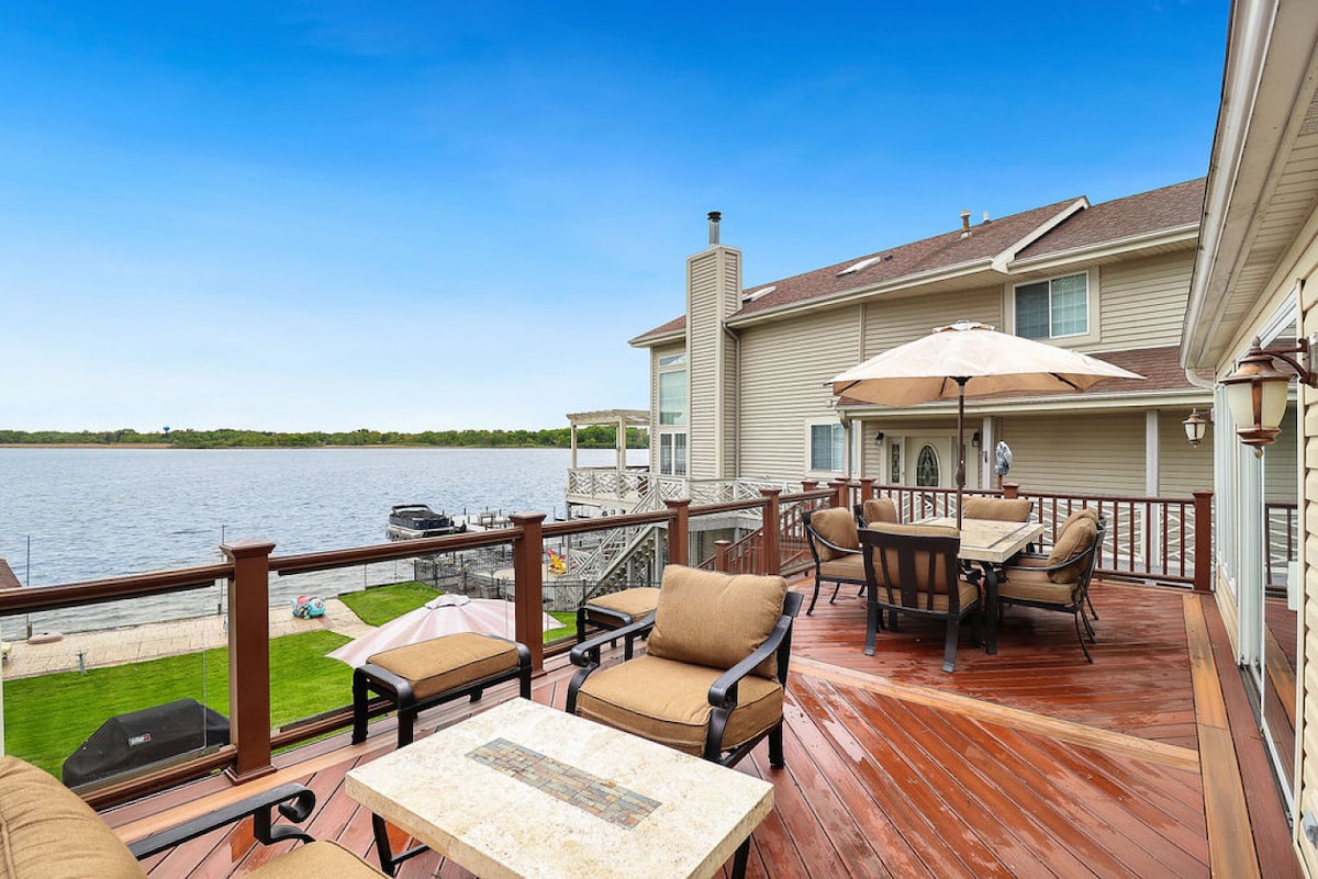 An outdoor deck features comfortable seating and a table with a fire pit. The lake is visible beyond the deck, with lawn chairs and umbrellas positioned on the grassy area at the water's edge. A pleasant view of the house can be seen in the background.