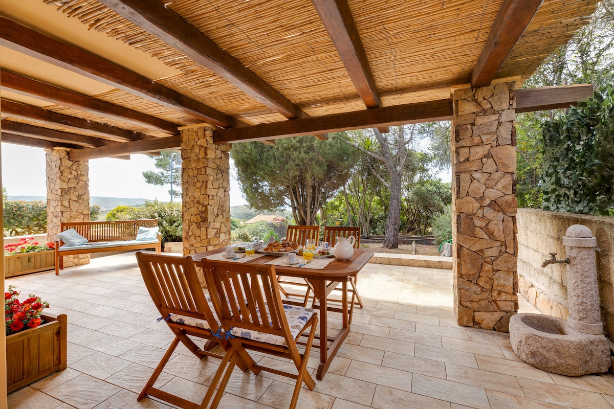 An outdoor veranda features wooden furniture, including a central dining table surrounded by chairs. Natural stone pillars support a textured ceiling, creating a shaded area. Lush greenery and blooming flowers provide a soft backdrop, enhancing the tranquil setting for outdoor relaxation.