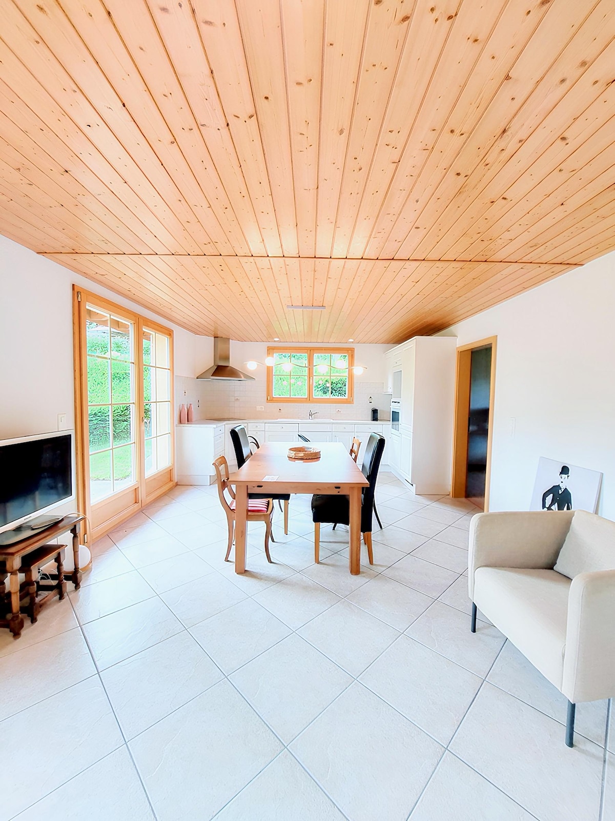 An open-plan living area features a wooden ceiling and tile flooring. A dining table with four chairs is centered, with a small television nearby. Large windows provide natural light and view of the greenery outside. A comfortable chair is positioned beside the table.