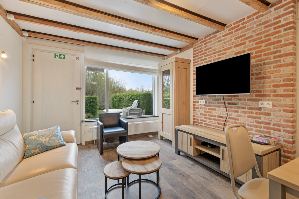 A living area features a light-colored sofa and a set of stacked circular coffee tables. A flat-screen TV is mounted on a brick wall. Large windows provide views of the outdoor greenery, allowing natural light to fill the space.