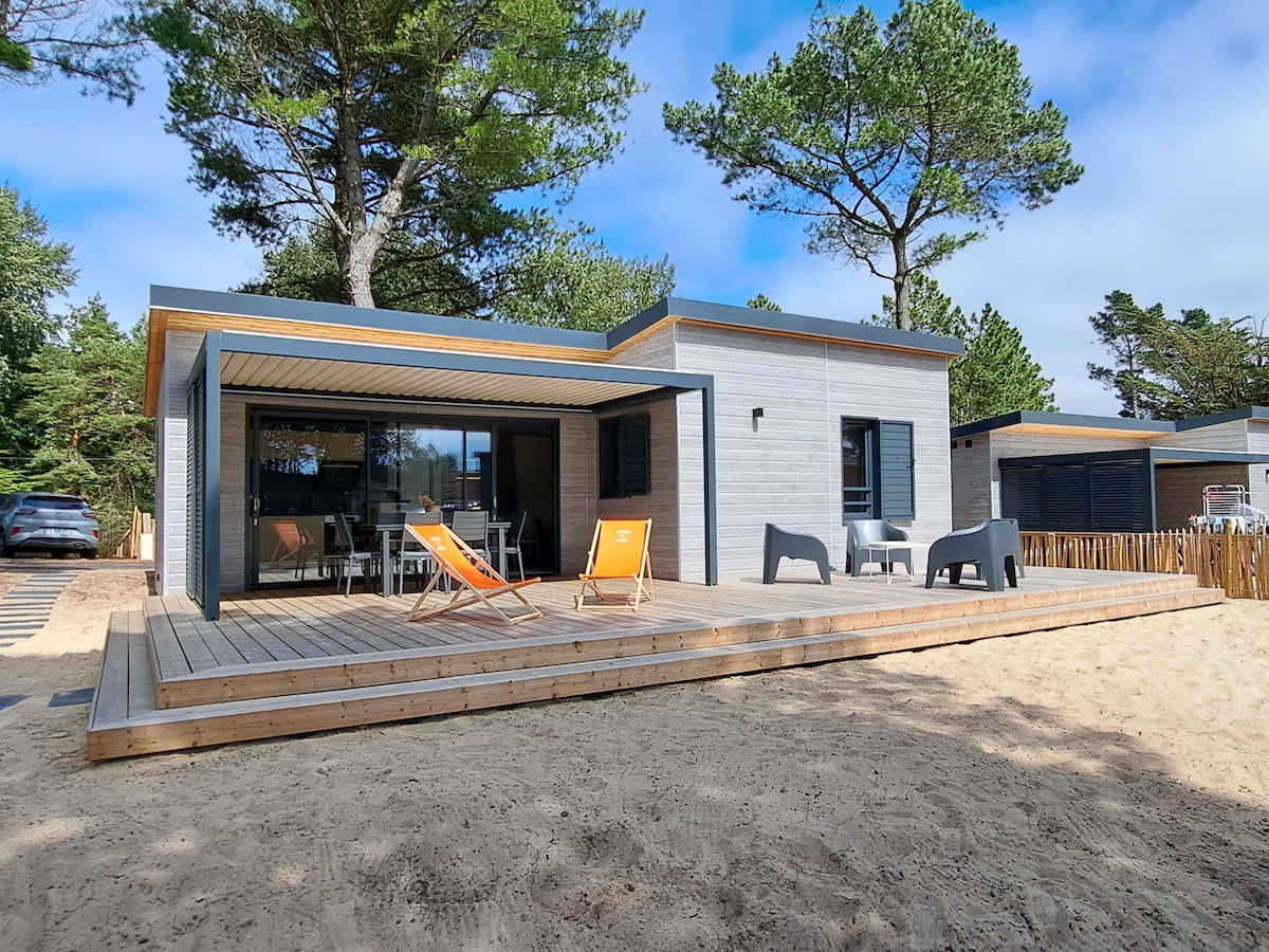 A modern chalet features a spacious wooden deck with orange and grey lounge chairs, positioned under tall trees. Large glass doors provide access to the interior, while a sandy area surrounds the outdoor space, enhancing the connection to nature.