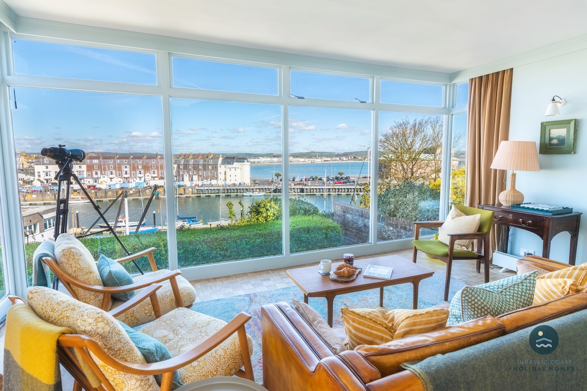 A spacious sitting room is framed by floor-to-ceiling glass windows, providing a clear view of Weymouth Harbour and the surrounding waterfront. Relaxed seating options are complemented by a telescope for viewing distant sights. Natural light enhances the room's bright and airy atmosphere.