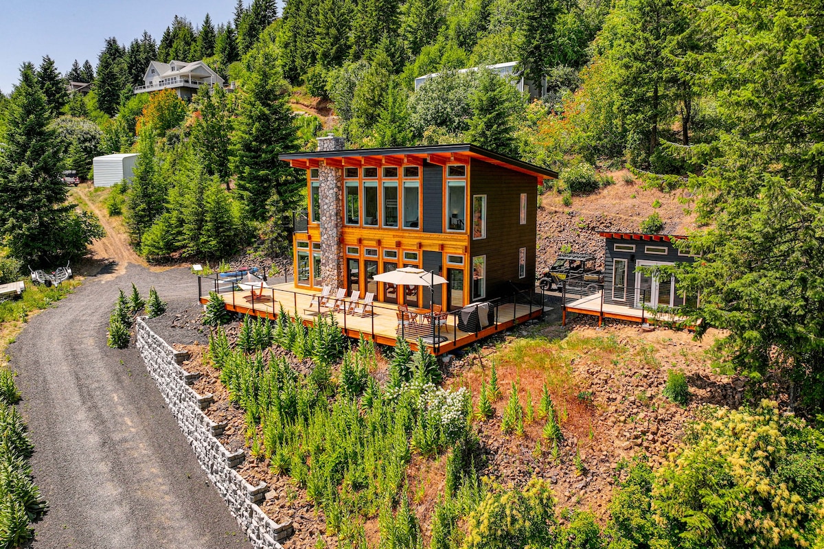 The modern lake house is set against a backdrop of trees and greenery. Large windows offer ample natural light, while the expansive deck is furnished with seating. The surrounding landscape features a gravel driveway and well-maintained shrubbery, contributing to a serene outdoor setting.