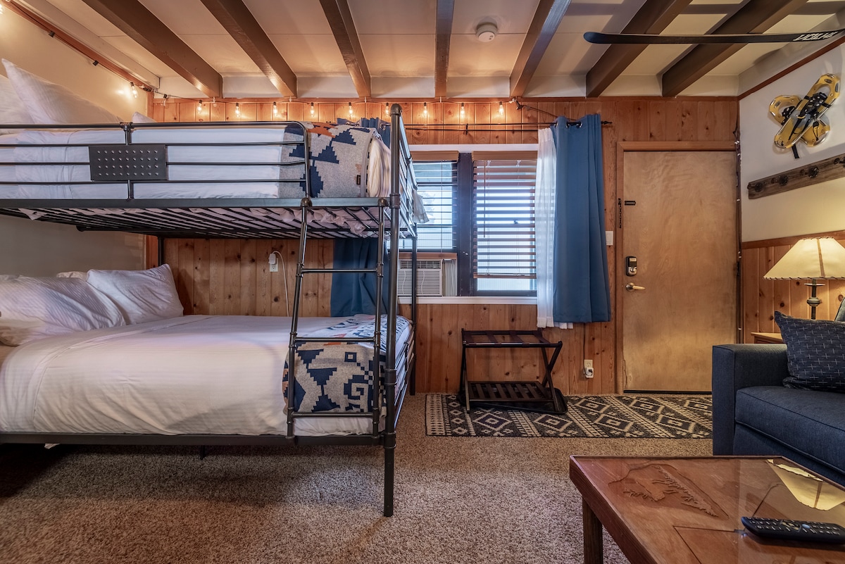 The studio features a bunk bed with a queen mattress on the bottom and a full mattress on the top. Soft beige carpeting complements the knotty pine paneling and eclectic decor. A window covered with light curtains adds natural light, while a sofa and coffee table provide additional seating.