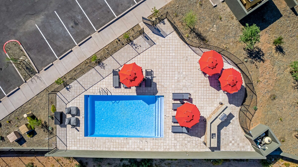 An aerial view captures a heated pool bordered by a spacious deck. Four bright red umbrellas offer shade for loungers positioned around the pool. The surrounding desert landscape is visible, along with a parking area in the background.