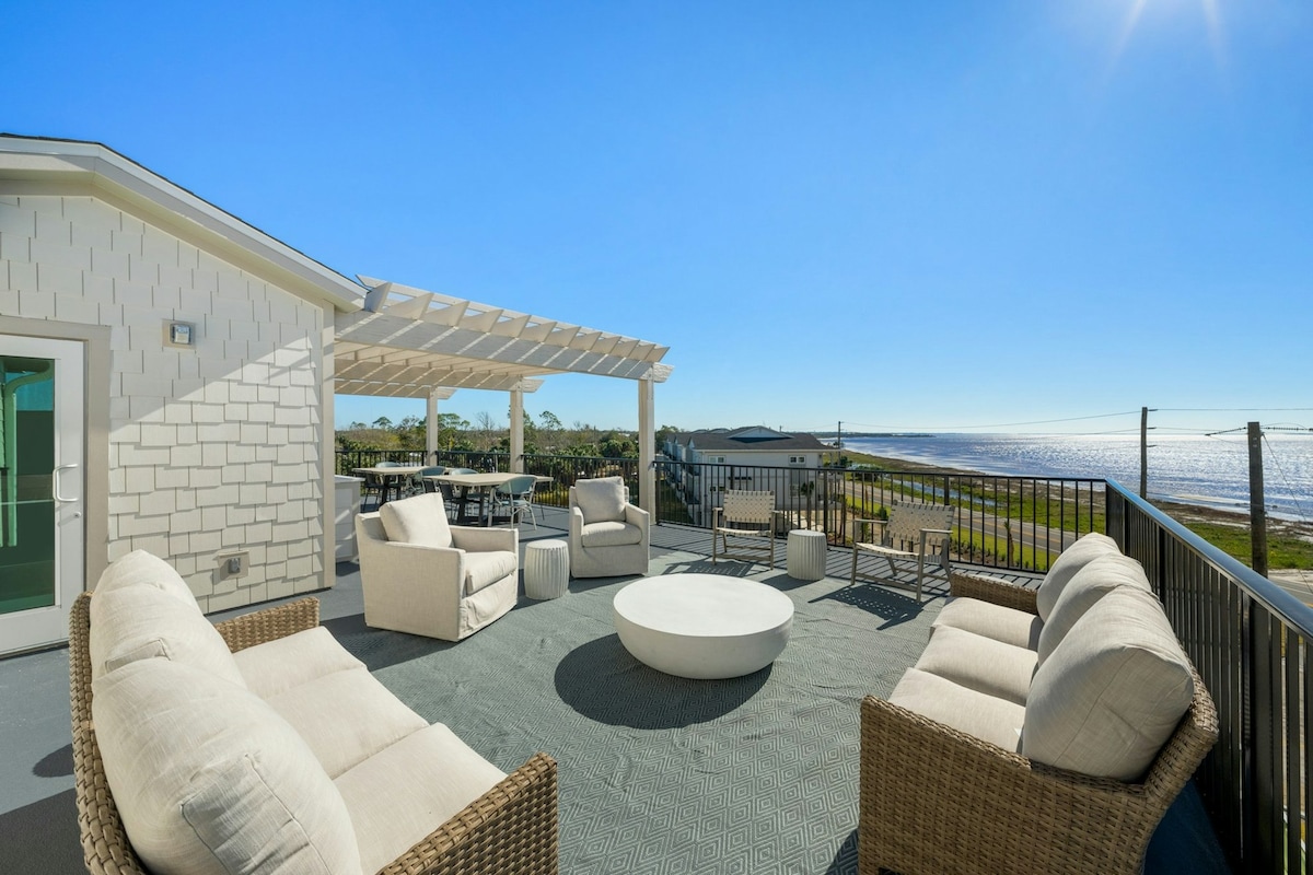 A spacious rooftop terrace features comfortable seating arranged around a central circular table. The area offers clear views of the ocean and surrounding landscape, with bright sunlight illuminating the space under a pergola.