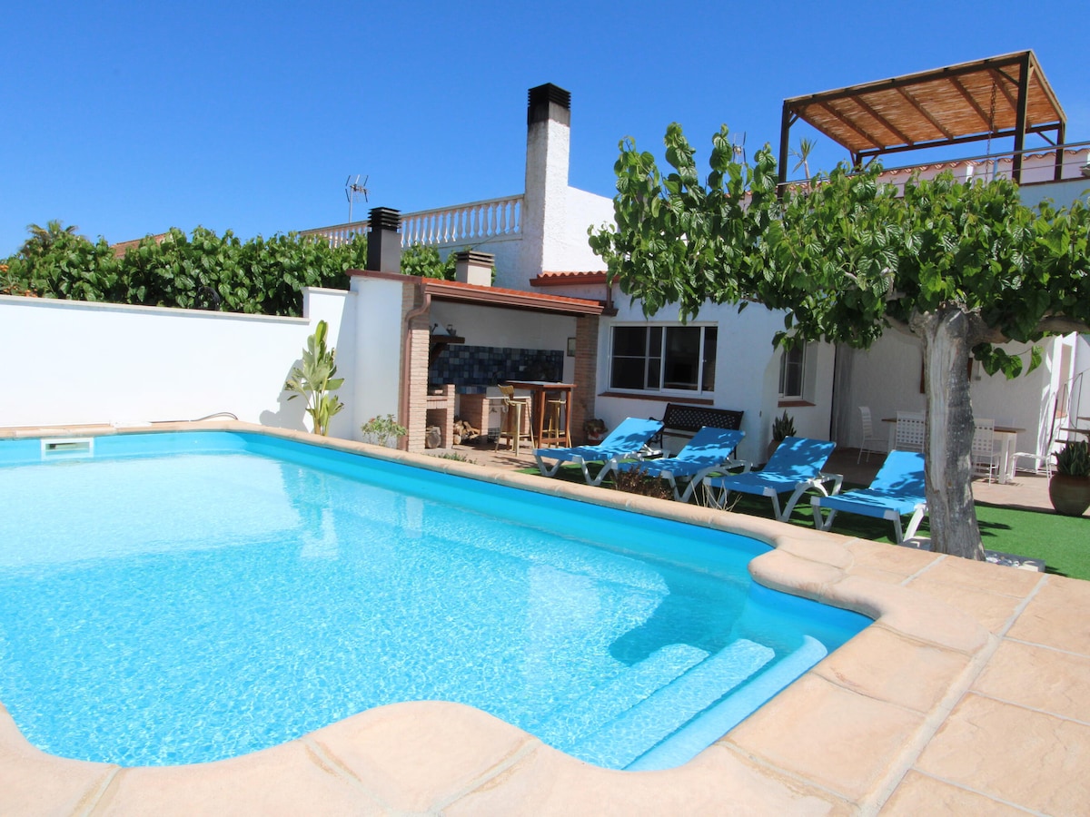 A private pool is shown surrounded by a spacious patio area. Blue lounge chairs are arranged nearby, with a barbecue area positioned in the background. A two-story structure is visible, complemented by lush greenery and a clear blue sky.