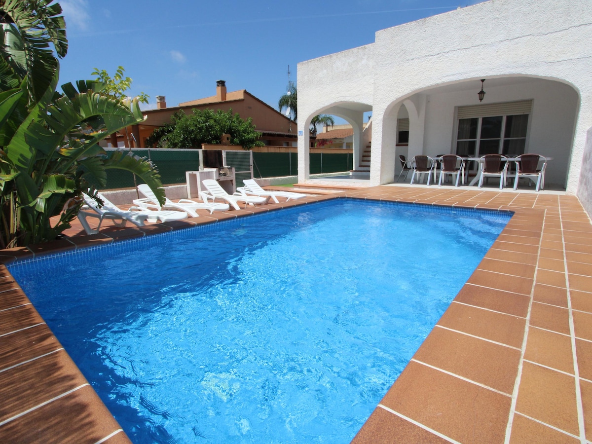 A private pool surrounded by a tiled patio offers space for relaxation. Lounge chairs are positioned nearby under the sun. An archway connects to an outdoor dining area, with a view of the pool and well-maintained landscaping in the background.