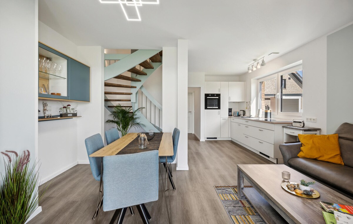 An open living space features a dining table surrounded by light blue chairs, positioned near a modern kitchen outfitted with ample cabinetry and appliances. A comfortable sofa with decorative cushions adds to the inviting atmosphere, while a staircase leads to an upper level.