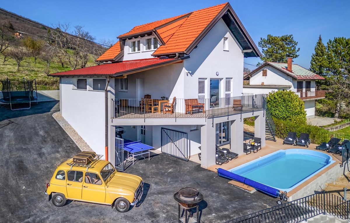 A two-story vacation home is showcased, featuring a warm orange-red roof and a light-colored exterior. A private pool sits in front, with seating areas and loungers nearby. A charming yellow car is parked in the foreground, and green hills provide a serene backdrop.