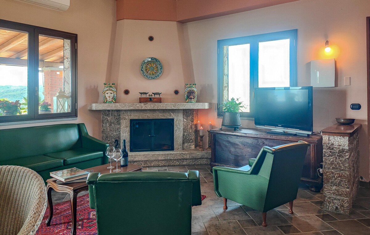 A cozy living area features comfortable green seating and a rustic stone fireplace. A wooden cabinet holds a television, and natural light enters through large windows. Decorative elements, including a wall clock and potted plants, enhance the inviting atmosphere.