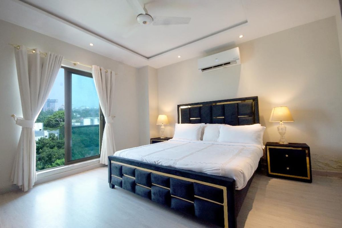 The bedroom features a spacious king-sized bed with a dark upholstered headboard, complemented by elegant bedside lamps. Large windows allow natural light to fill the room, offering views of the greenery outside. Soft, neutral tones create a calming atmosphere.