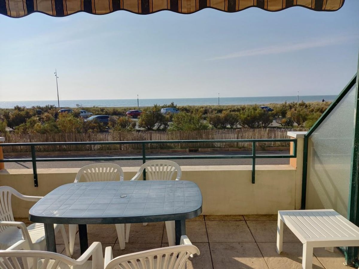 A spacious balcony is equipped with a large rectangular table and several white chairs, offering a view of the sea. The setting captures a coastal atmosphere, with vegetation and parked vehicles visible in the foreground, enhancing the outdoor experience.