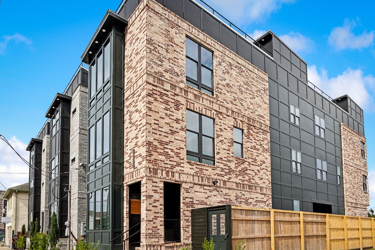 A modern multi-story building features a combination of brick and sleek black paneling. Large windows are prominently displayed, allowing ample natural light. A fenced area surrounds the property, enhancing privacy, while the contemporary design integrates seamlessly into the neighborhood.