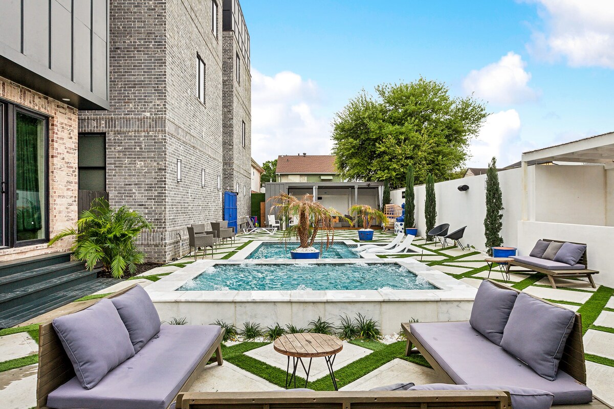 An outdoor space features a small pool surrounded by landscaped areas with green grass. Comfortable seating areas with gray cushions are positioned nearby. The walls of the building are made of light-colored brick, and tall plants provide a touch of greenery.