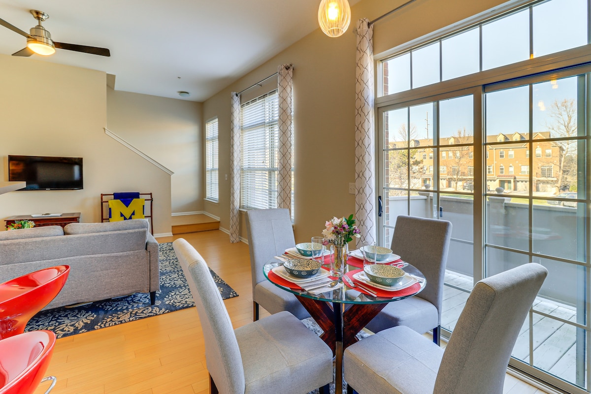 A bright living area with large windows showcases a comfortable seating arrangement and a smart TV. A dining table for four is elegantly set, surrounded by upholstered chairs. Light filters through sheer curtains, reflecting off bamboo flooring that extends towards the staircase.