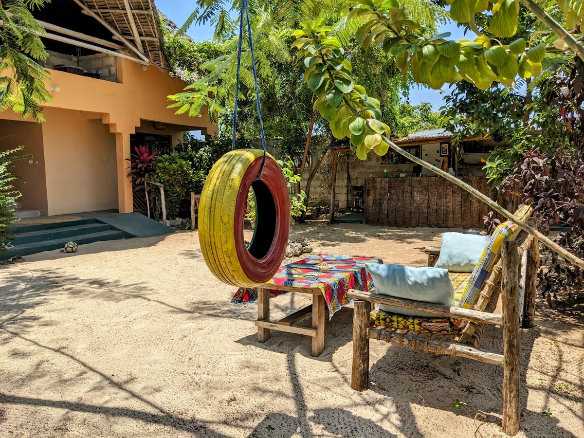 A private garden space is featured, showcasing a colorful seating area with a patterned fabric draped over a wooden table. A yellow tire swing is suspended from a lush tree branch, surrounded by greenery and sandy ground, contributing to a relaxed atmosphere.