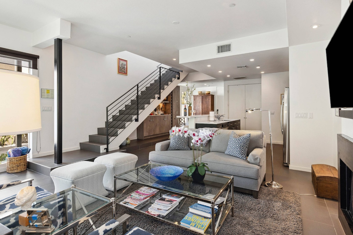 An open-concept living area features a plush gray sofa adorned with decorative pillows, complemented by a glass coffee table with magazines. The space is bathed in natural light, highlighting modern furnishings and a staircase leading to the upper level. A dining area and kitchen are visible in the background.
