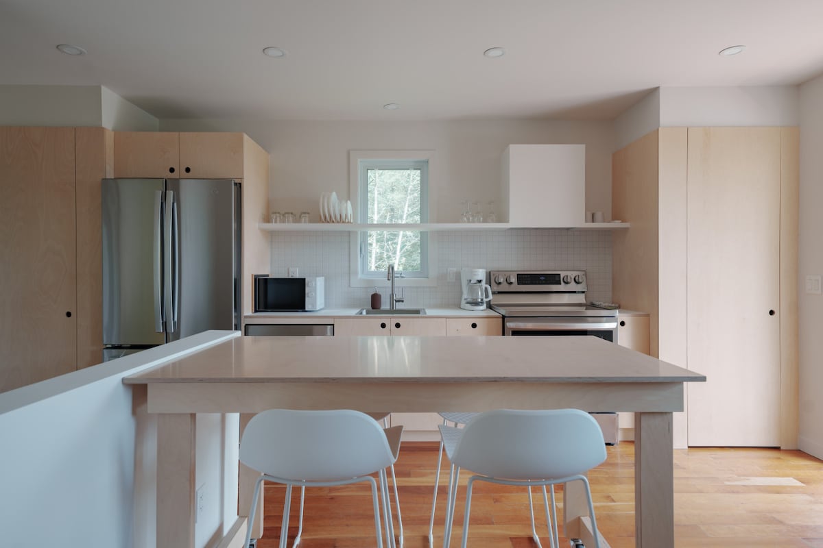 A modern kitchen features light wood cabinetry and a central dining table with white chairs. Stainless steel appliances, including a refrigerator and oven, are visible, along with a microwave. Natural light enters through a window, enhancing the inviting environment.