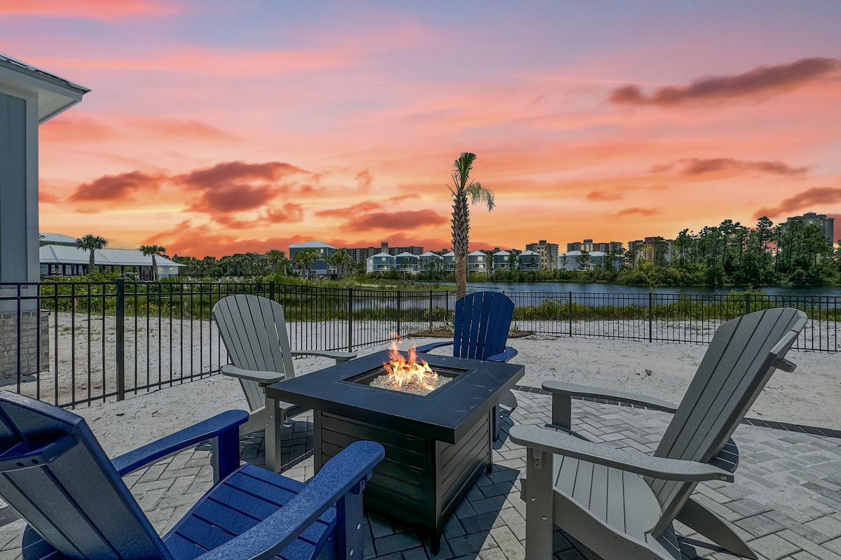 A cozy outdoor seating area features four adirondack chairs arranged around a fire pit, set against a vibrant sunset. The foreground shows a sandy area, with a calm lake and distant buildings visible in the background, creating a serene atmosphere.