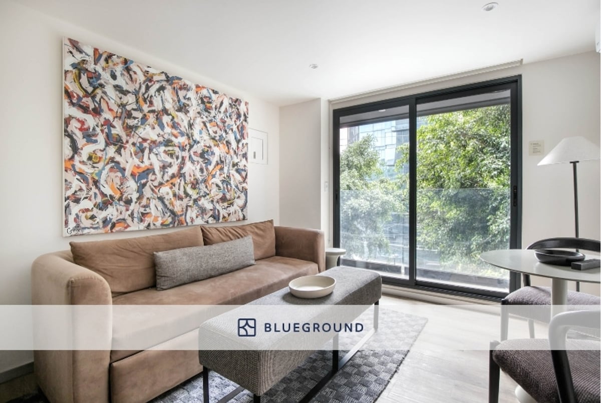 A light-filled living area features a comfortable brown sofa with decorative pillows, set against a colorful wall art piece. A round coffee table is placed in front of the sofa, while a large window showcases views of greenery outside.