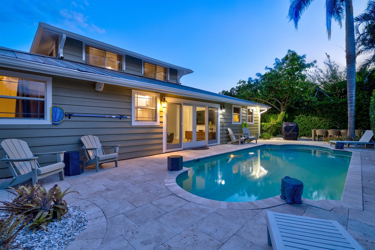 A serene outdoor space features a private heated pool surrounded by tropical greenery. Lounge chairs are positioned near the pool, while the two-story home showcases glass sliding doors, inviting natural light and creating an open atmosphere for relaxation.