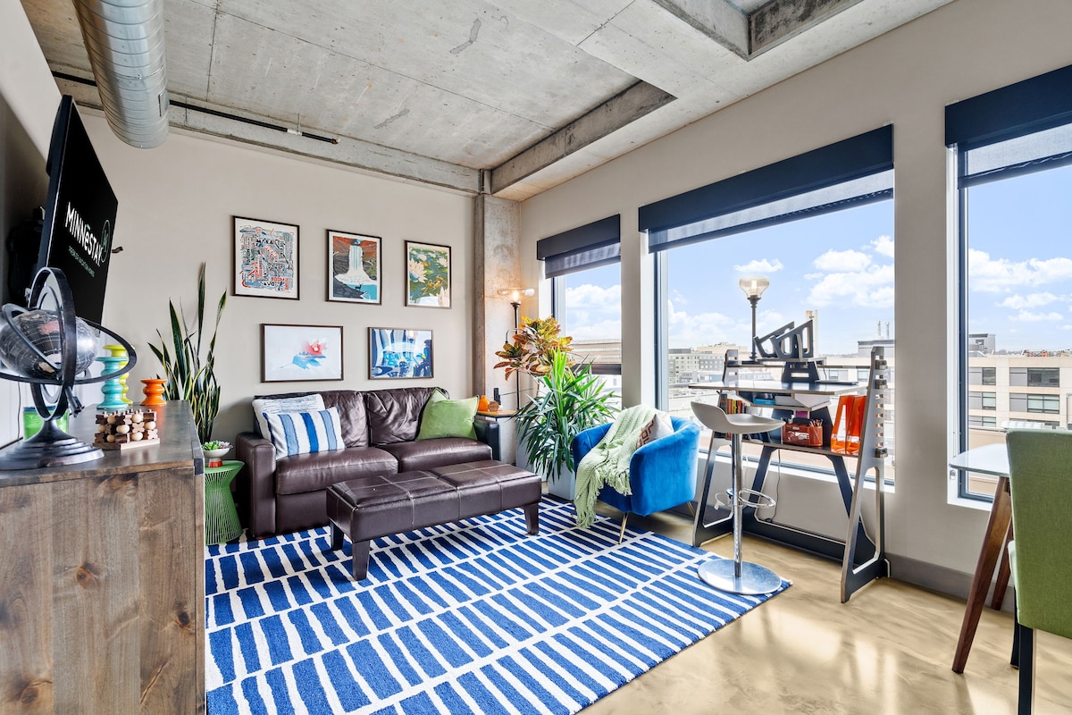 A modern living area features a leather couch paired with colorful accent chairs. A striped area rug adds texture, while large windows frame city views. Artwork hangs on the walls, and a desk beside the window offers a workspace with natural light.