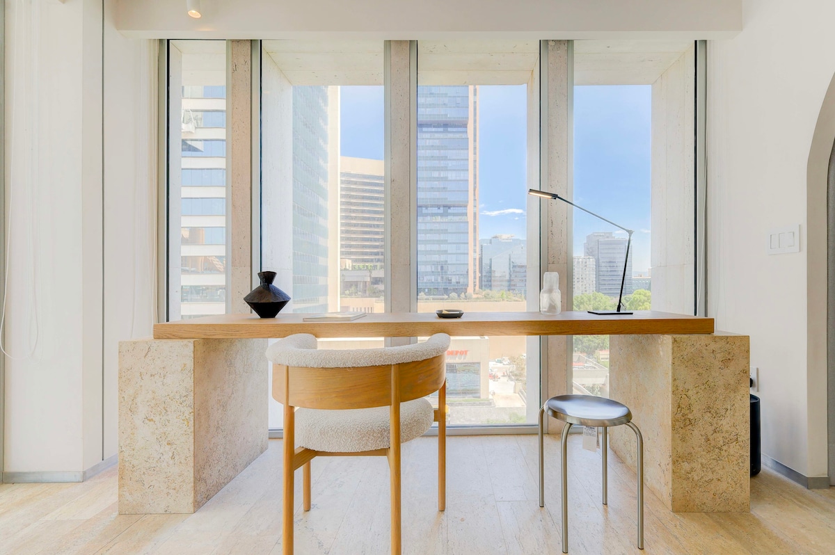 A serene work area features a wooden desk set against expansive floor-to-ceiling windows, offering views of the cityscape. A minimalist chair complements the decor, with a decorative vase and a lamp placed thoughtfully on the desk.