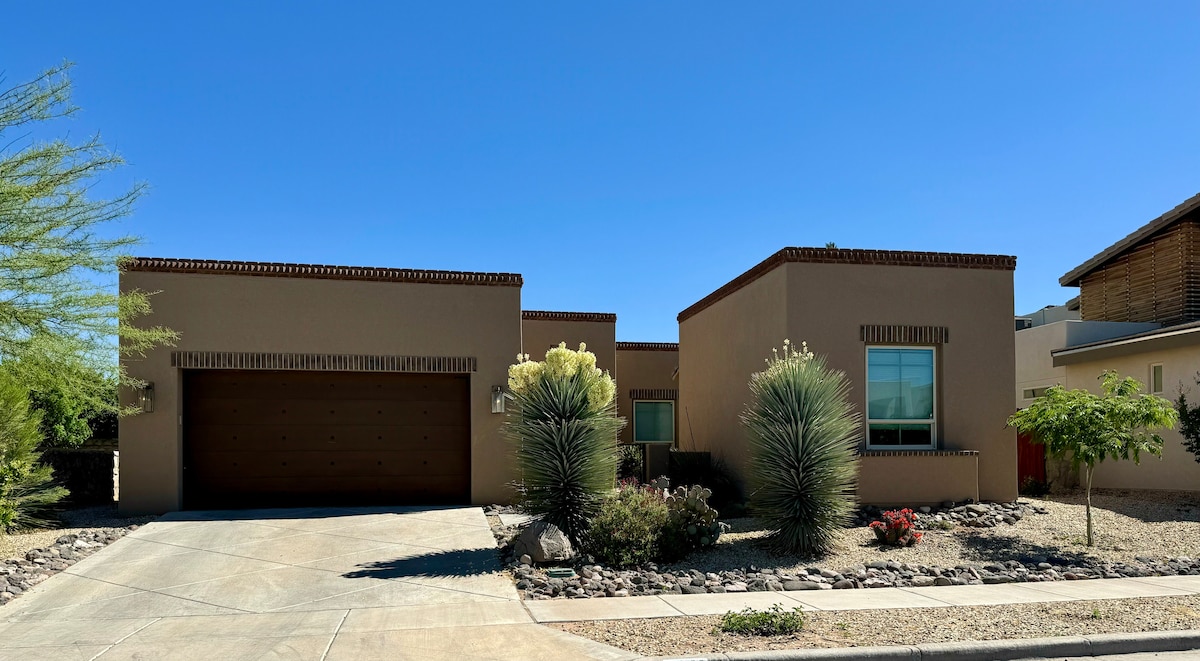 A modern single-story house featuring a two-car garage. A well-kept landscape includes desert plants and low-maintenance ground cover. Large windows provide natural light, and the home’s warm tones complement the clear blue sky.