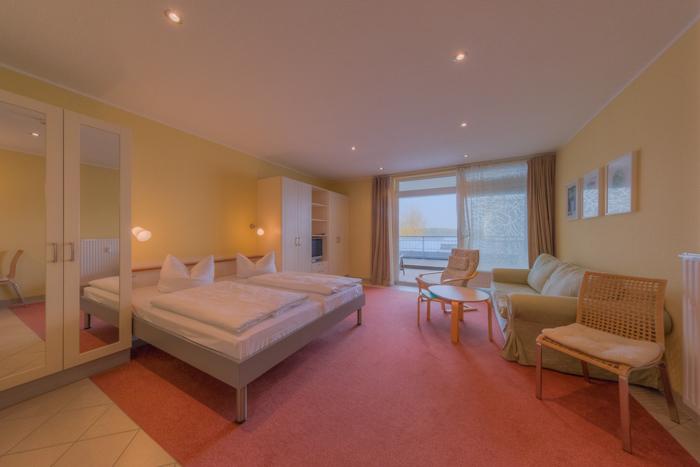 A spacious living area is illuminated by natural light coming through large windows. The room features two separate beds with fresh linens, a comfortable seating area, and a small dining table. A wardrobe is visible, providing ample storage space.